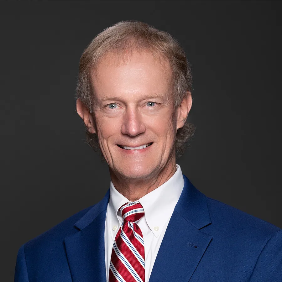 Smiling middle-aged man in blue suit jacket, white shirt, and red striped tie against a dark background.