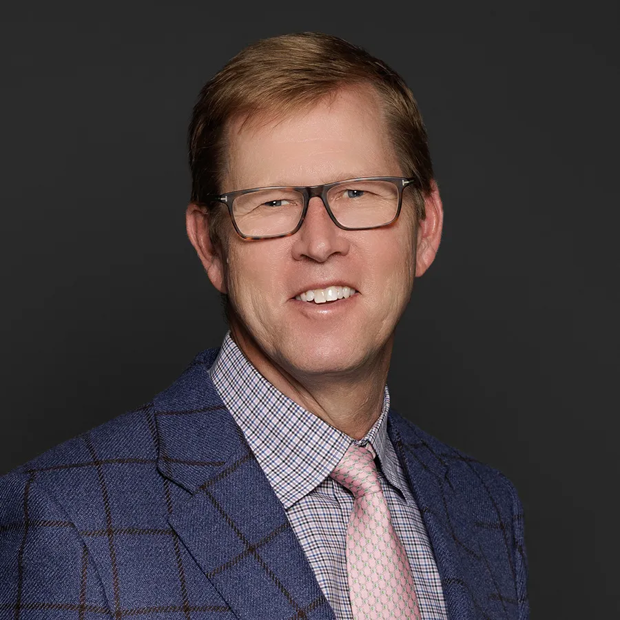 Middle-aged man with glasses wearing a blue checked blazer, plaid shirt, and pink tie smiling against black background