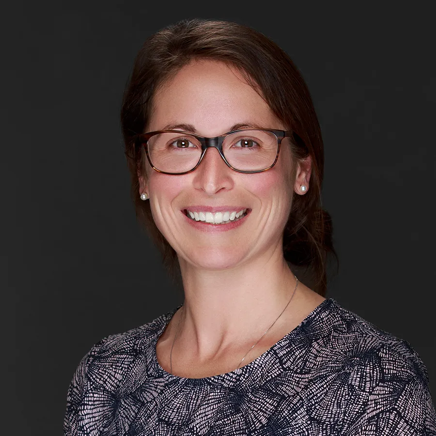 Smiling woman wearing glasses and patterned top against a plain dark background.