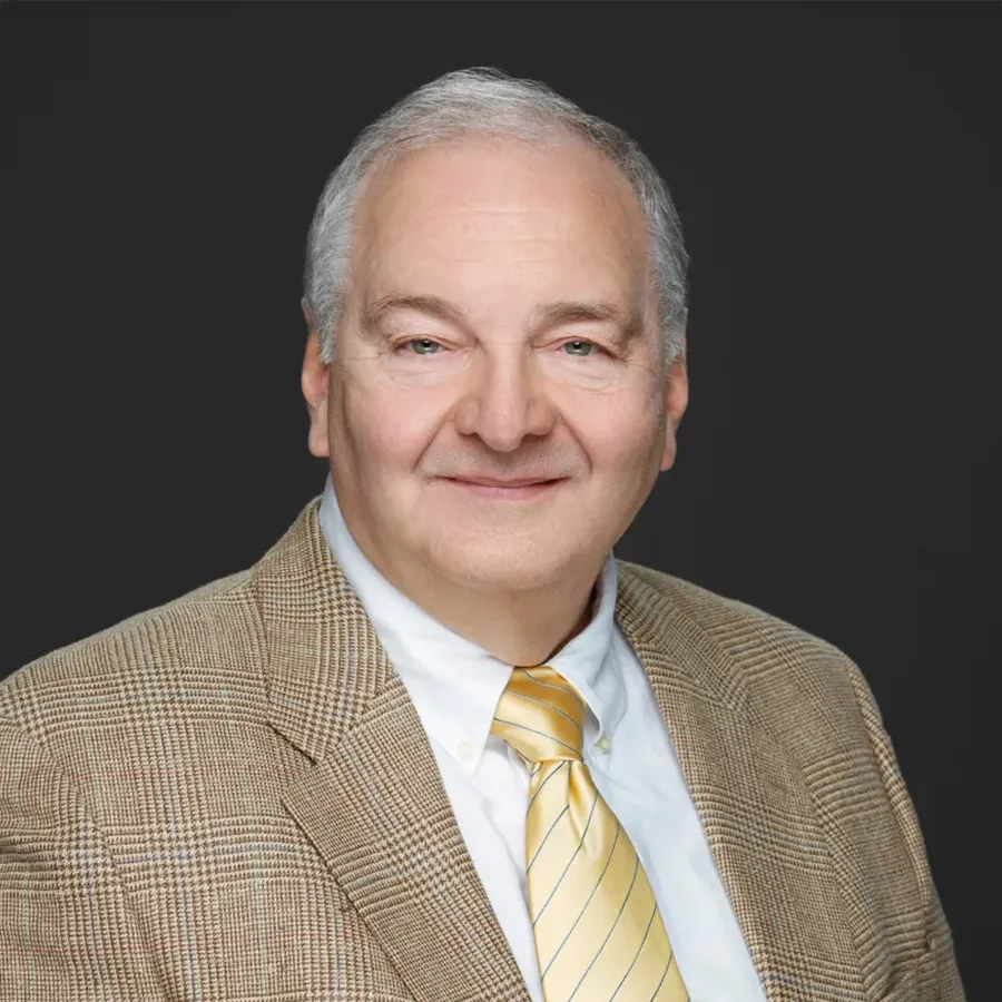 Portrait of older man with gray hair in checkered blazer and yellow tie smiling against dark background