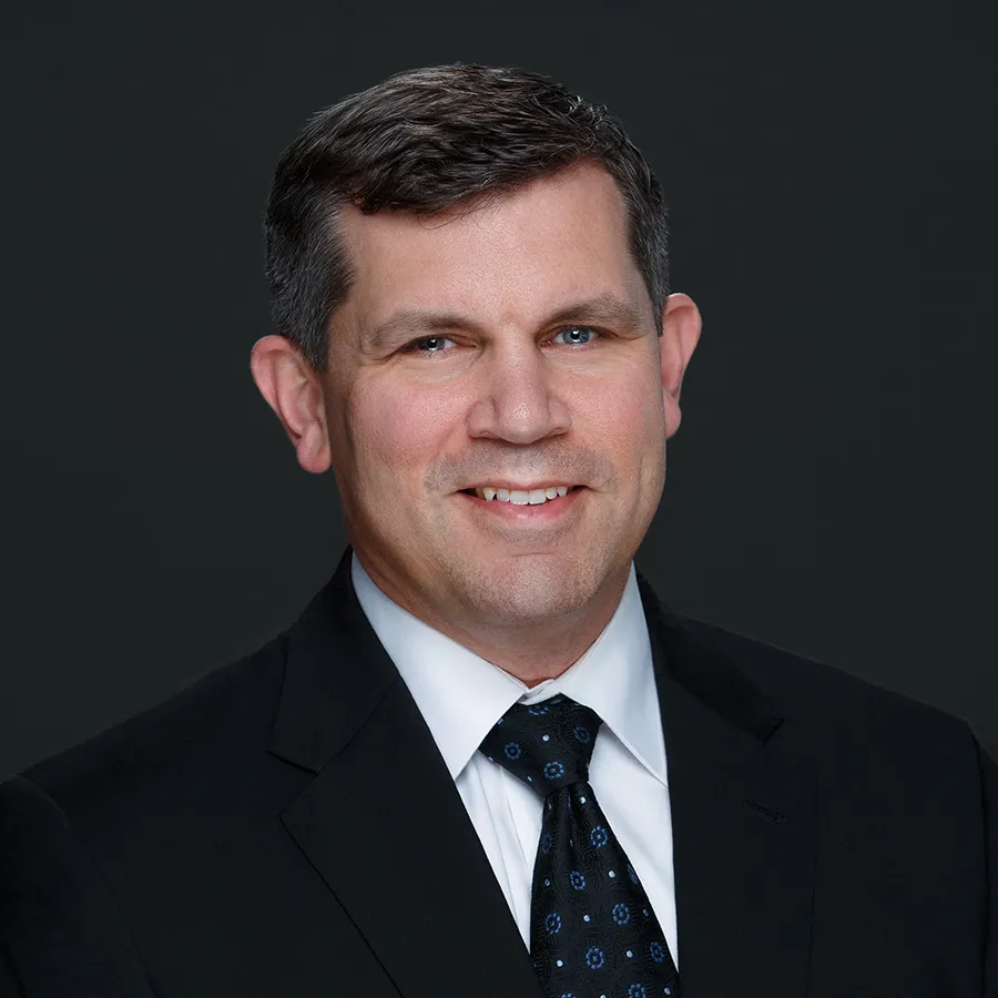 Professional headshot of a smiling middle-aged man in a black suit with patterned tie against dark background