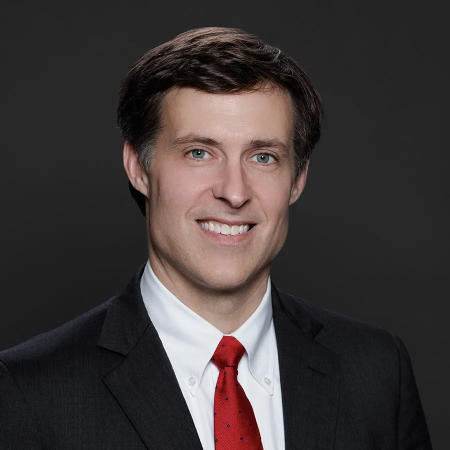 Professional man in dark suit, white shirt, and red tie smiling against a dark background