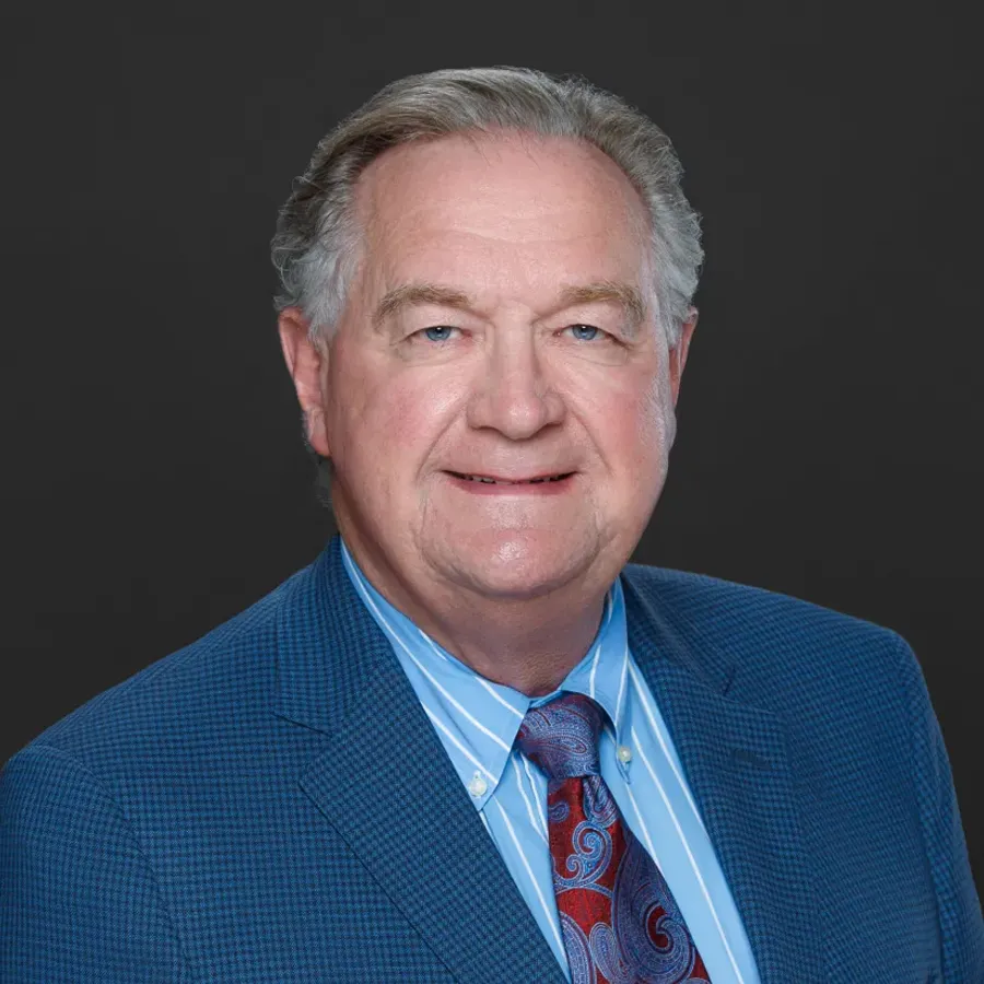 Older man with gray hair wearing a blue checkered suit, striped shirt, and paisley tie smiling against dark background