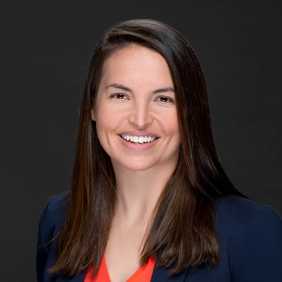 Smiling woman with long brown hair wearing a navy blazer and red top against a dark background.
