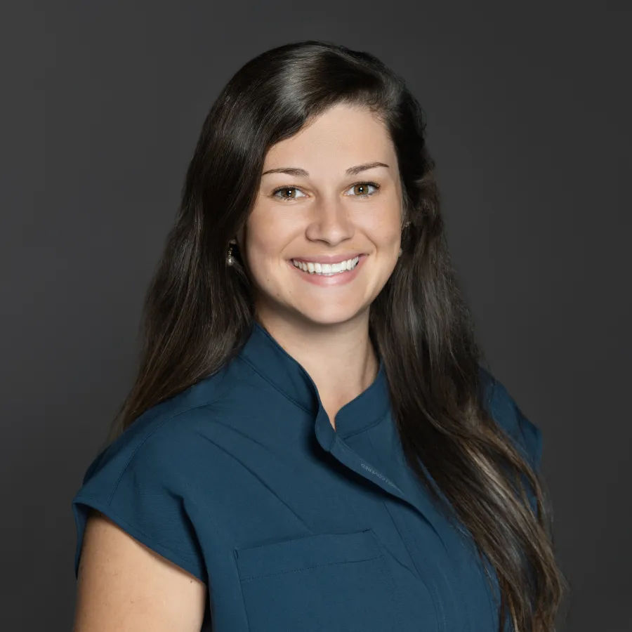 Smiling woman with long brown hair wearing a blue sleeveless top against a dark gray background.