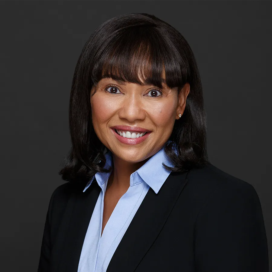 Smiling professional woman with dark hair wearing a black blazer and light blue collared shirt on gray background