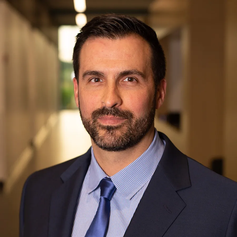 Professional man with beard wearing a navy suit and blue tie standing in a modern office hallway.