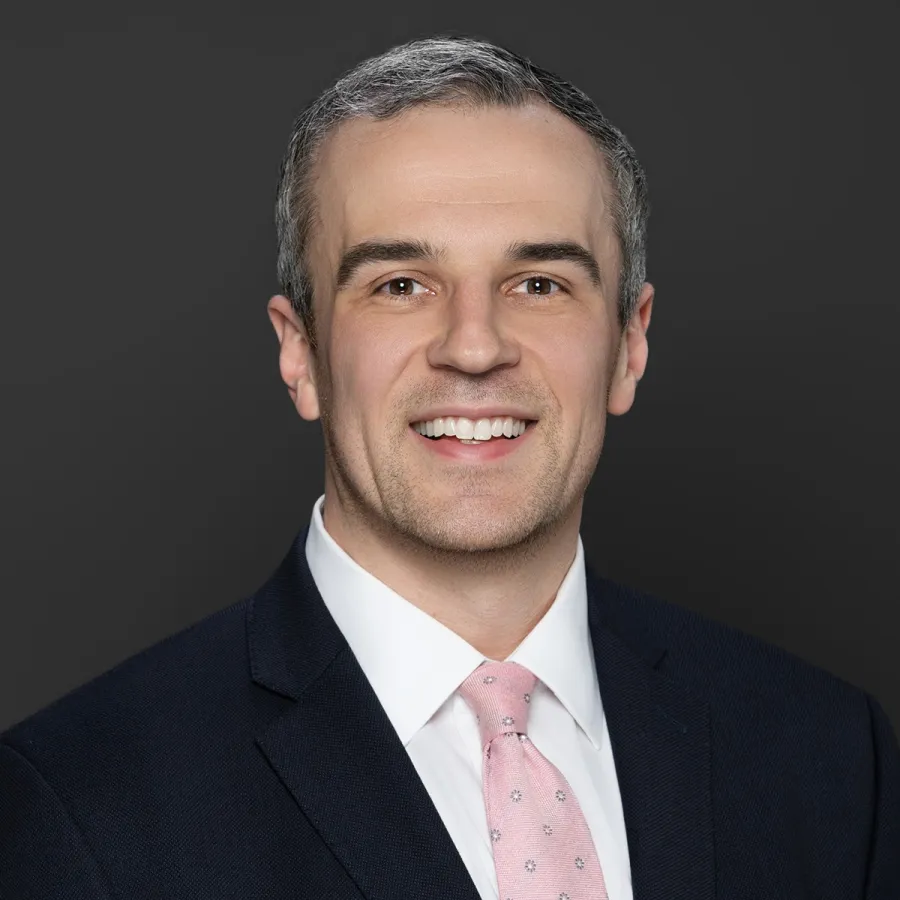 Smiling man in a dark suit jacket, white shirt, and light pink patterned tie against a dark background.