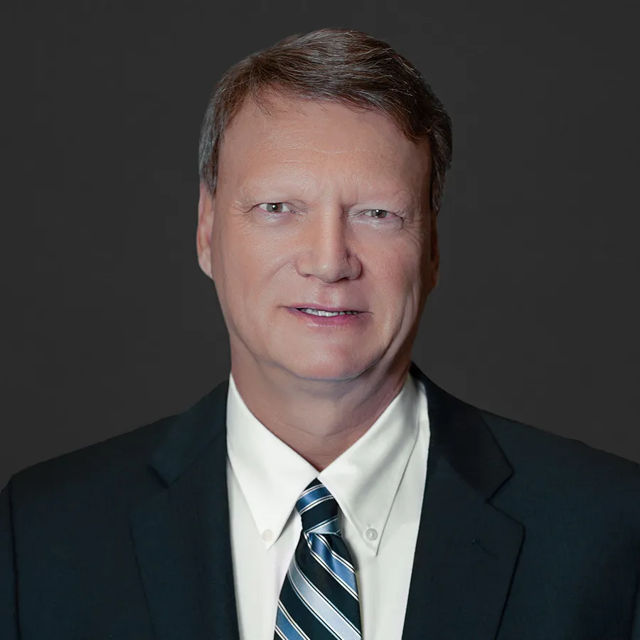 Professional middle-aged man wearing a dark suit, white shirt, and striped tie against a dark background