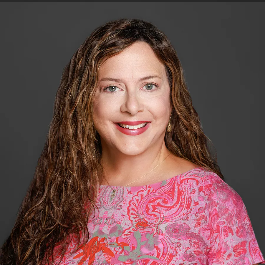 Smiling woman with long curly brown hair wearing a pink patterned blouse against a gray background