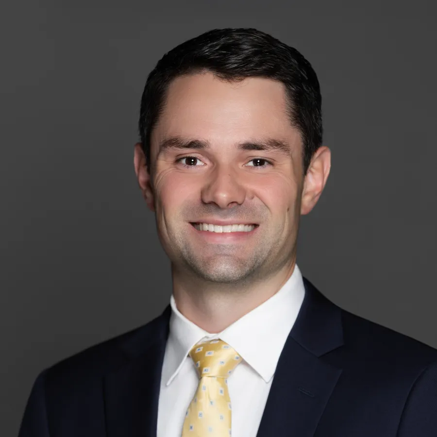 Professional headshot of a smiling man wearing a navy suit and yellow patterned tie against a gray background