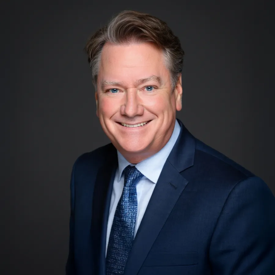 Smiling middle-aged man in a navy blue suit and patterned tie against a dark background.