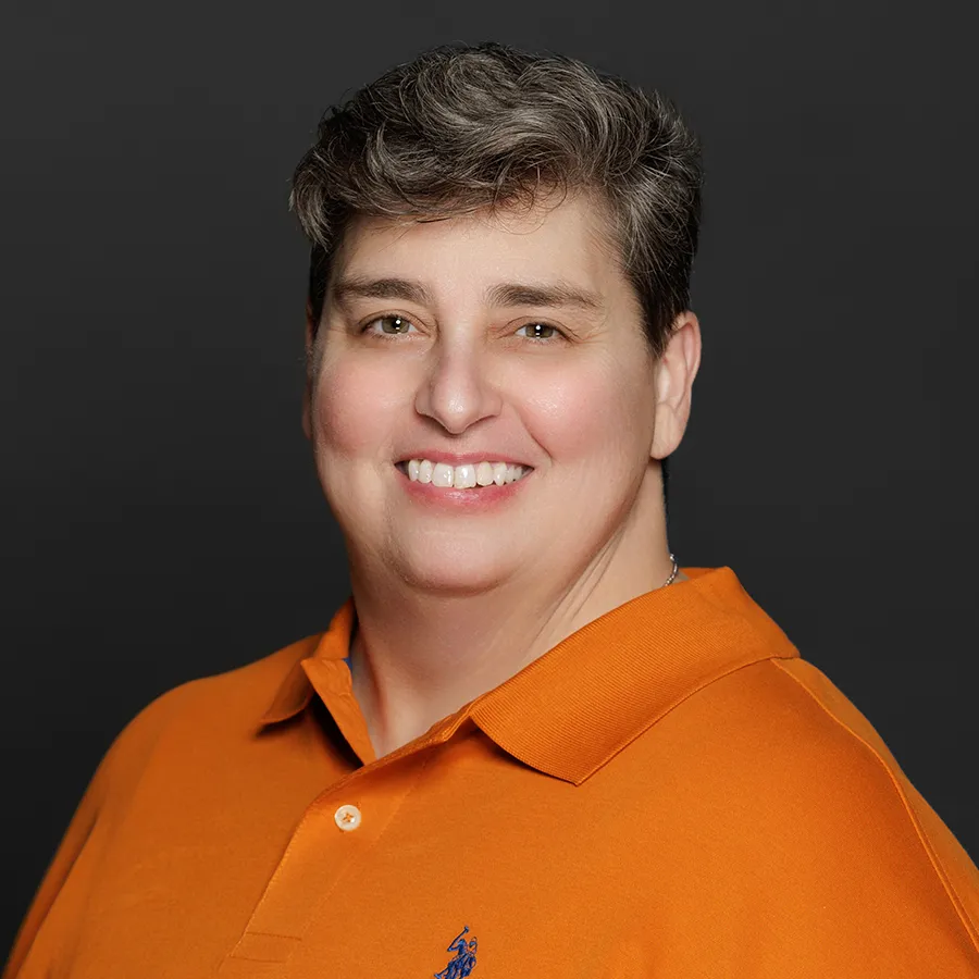 Smiling person with short gray hair wearing an orange polo shirt against a dark background