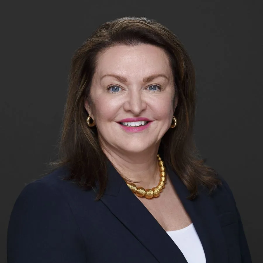 Professional headshot of smiling woman with brown hair wearing gold necklace, earrings, and navy blazer on gray background