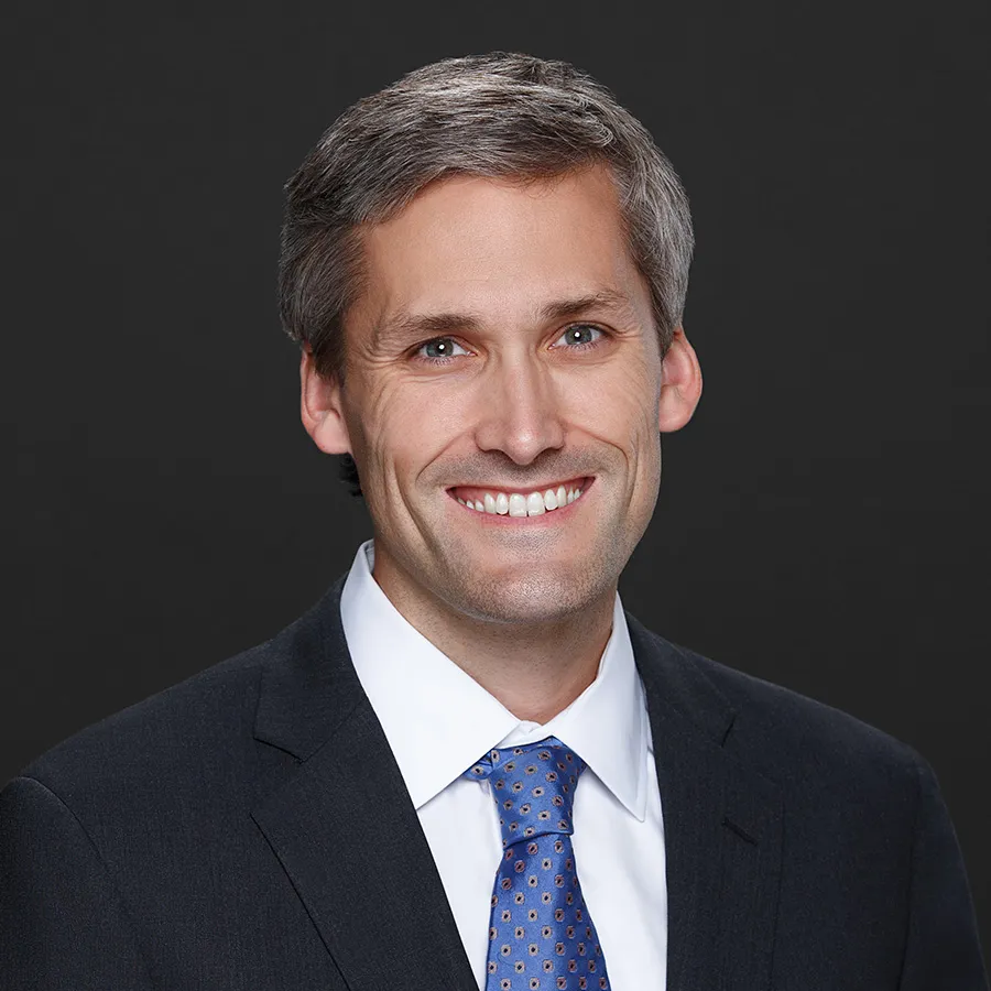 Smiling middle-aged man in a dark suit and blue patterned tie with a white shirt against a black background.