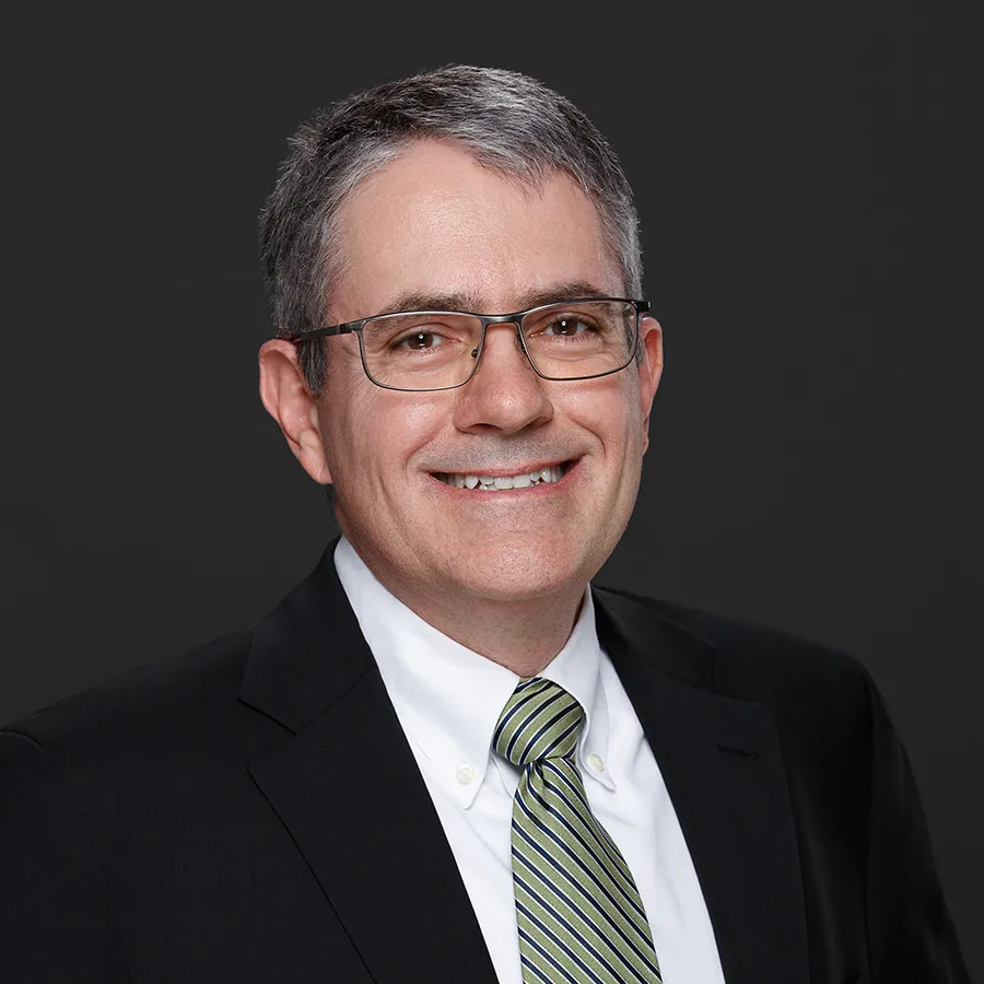Middle-aged man wearing glasses, a black suit, white shirt, and green striped tie smiling against a dark background