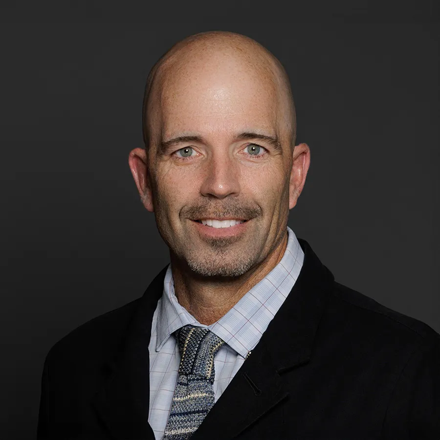 Portrait of a smiling bald man in a suit and patterned tie against a dark gray background