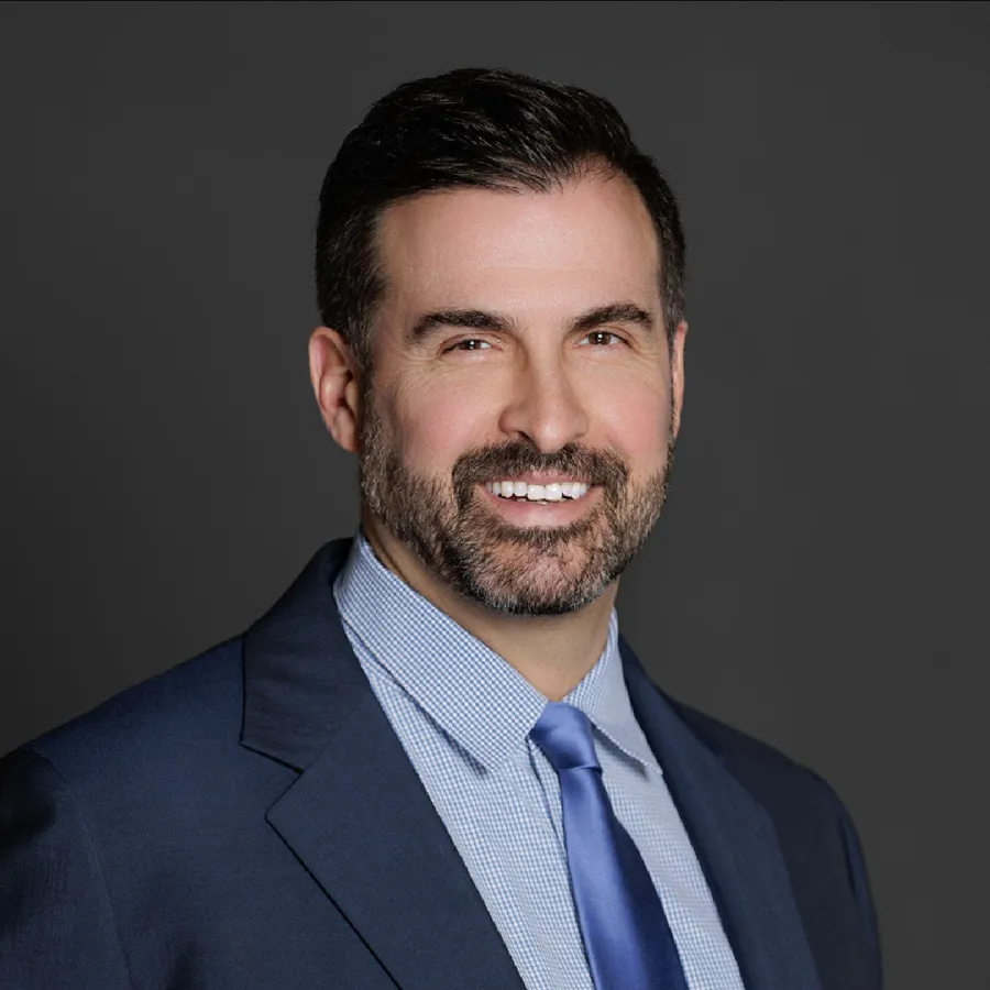 Smiling man in a navy suit, light blue shirt, and blue tie against a dark gray background.