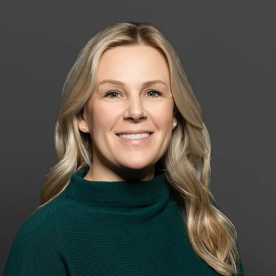 Smiling blonde woman with long hair wearing a dark green sweater against a gray background