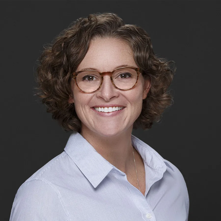 Smiling woman with short curly hair and glasses wearing a light blue striped shirt against a dark background