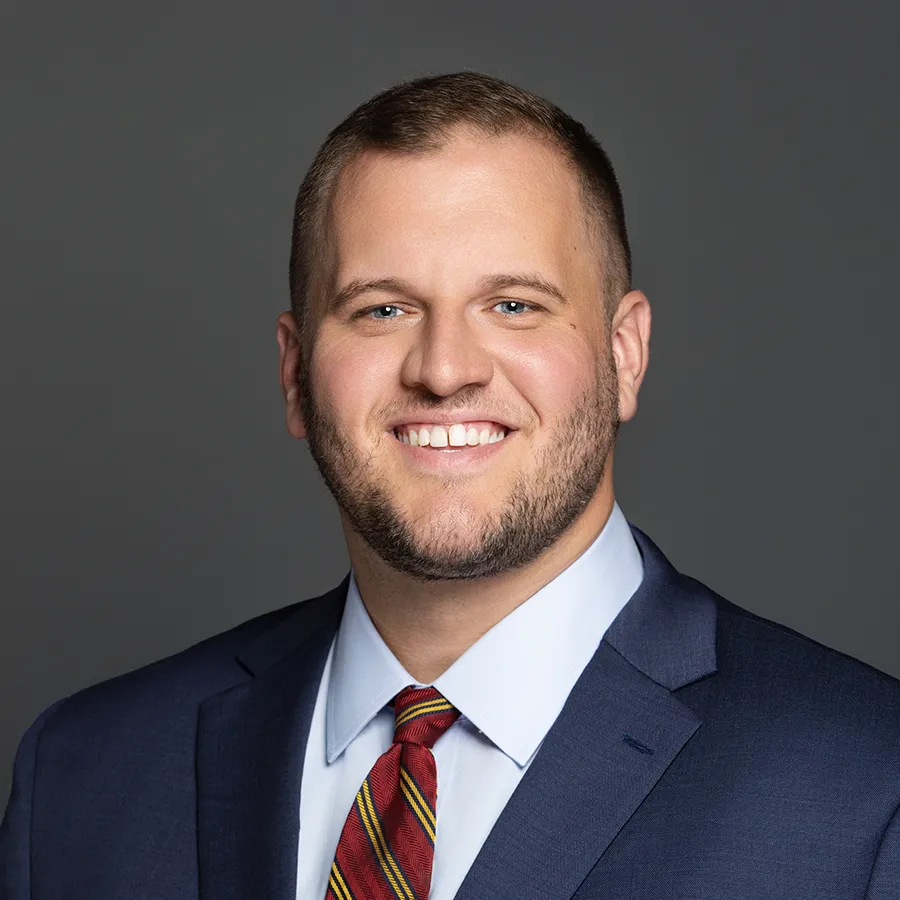 Smiling man with short hair, beard, and blue eyes wearing a navy suit, white shirt, and striped red tie.