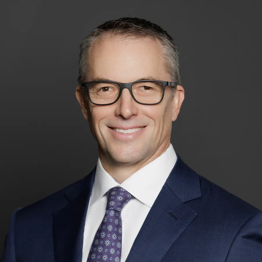 Professional middle-aged man in glasses wearing navy blue suit and patterned tie smiling against gray background