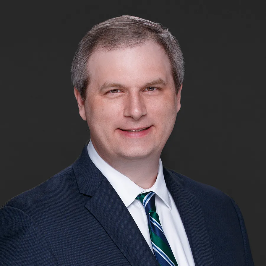 Professional headshot of a middle-aged man in a navy suit, white shirt, and striped blue and green tie against a dark background