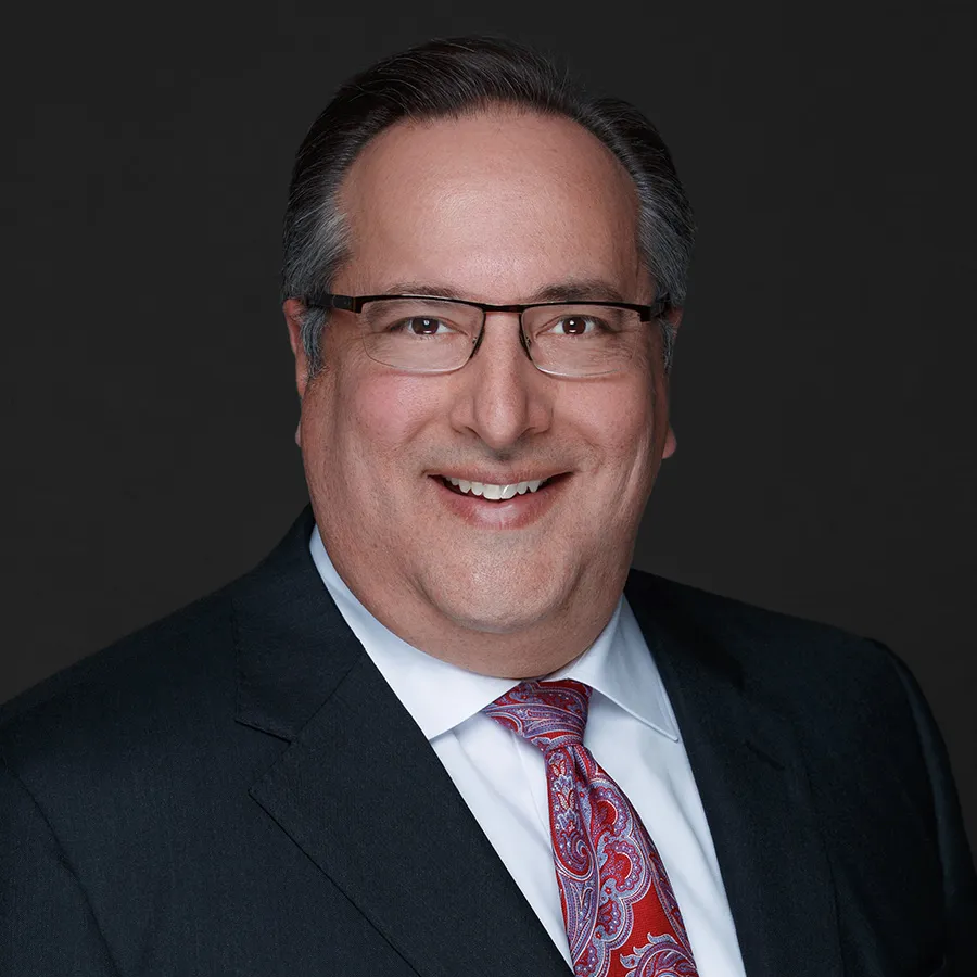 Smiling middle-aged man wearing glasses, dark suit, white shirt, and red paisley tie against dark background