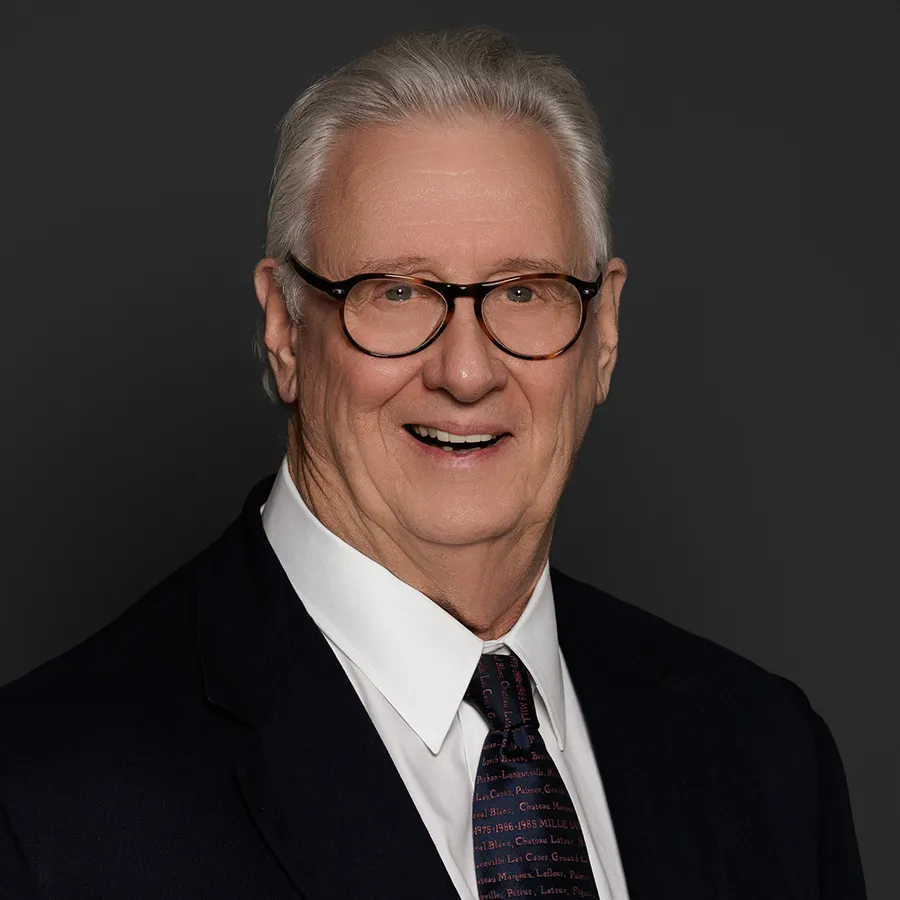 Elderly man with silver hair and glasses wearing a dark suit, white shirt, and patterned tie against black background