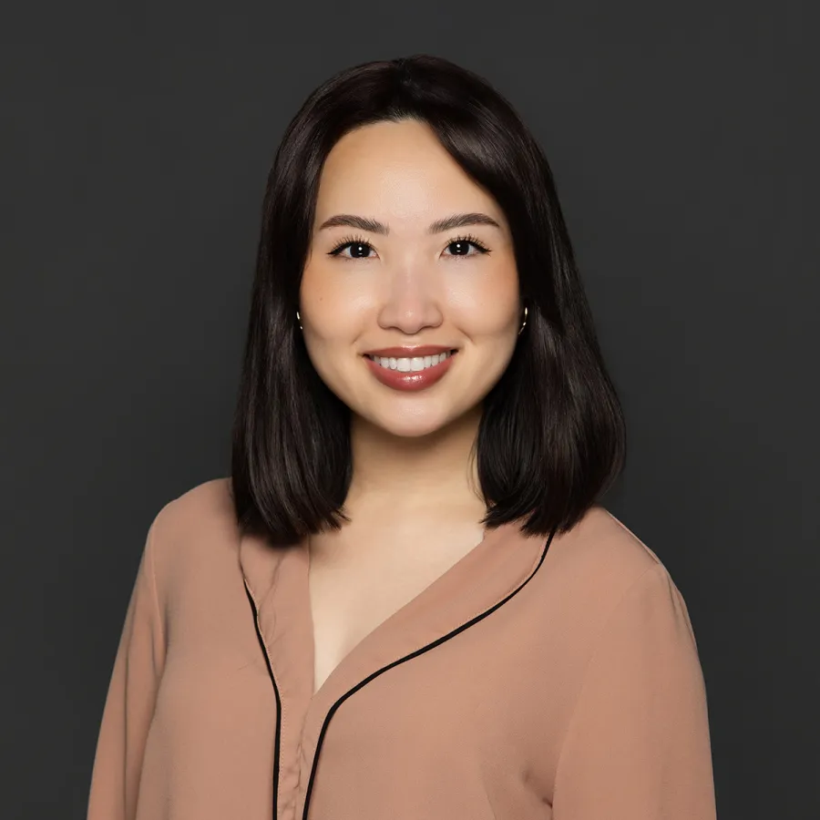 Young woman with straight dark hair smiling, wearing a beige blouse with black trim against a dark background