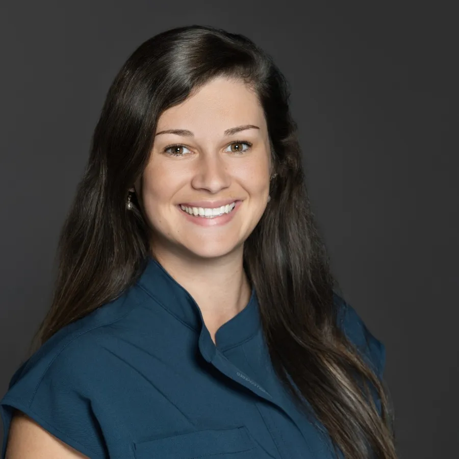 Smiling woman with long brown hair wearing a blue sleeveless top against a dark gray background.