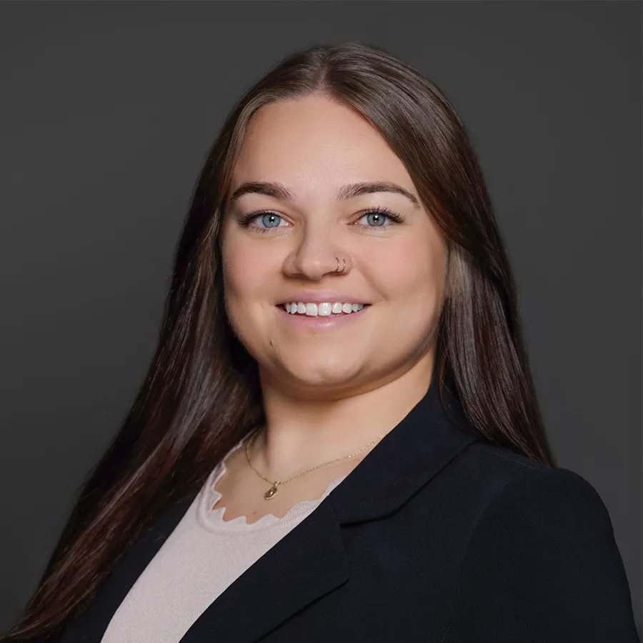 Smiling young woman with long brown hair wearing a black blazer and light top against a gray background