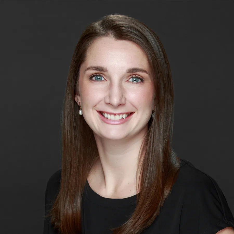 Smiling woman with straight brown hair wearing pearl earrings and black top against dark background