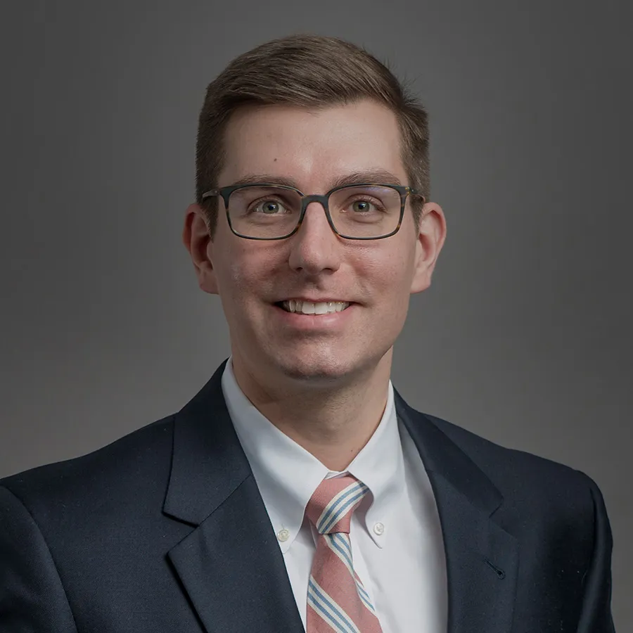 Professional man wearing glasses, a dark suit, white shirt, and striped tie smiling against gray background