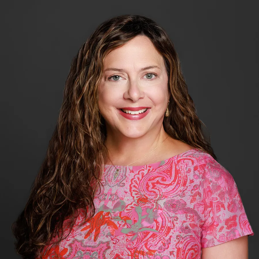 A smiling woman with long, curly hair wearing a colorful floral top against a dark background.