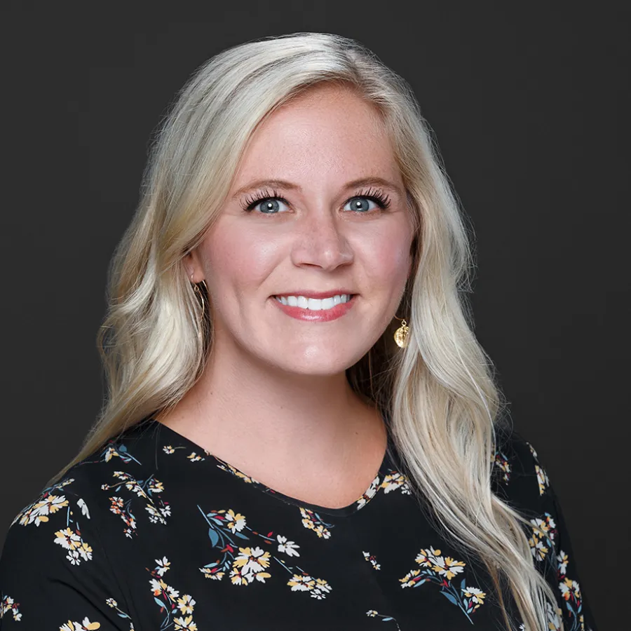 Smiling blonde woman with long hair wearing floral black top and gold earrings against a dark background