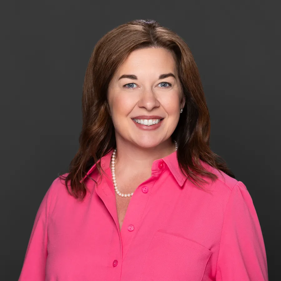Smiling woman with brown hair wearing a bright pink blouse and pearl necklace on a dark gray background