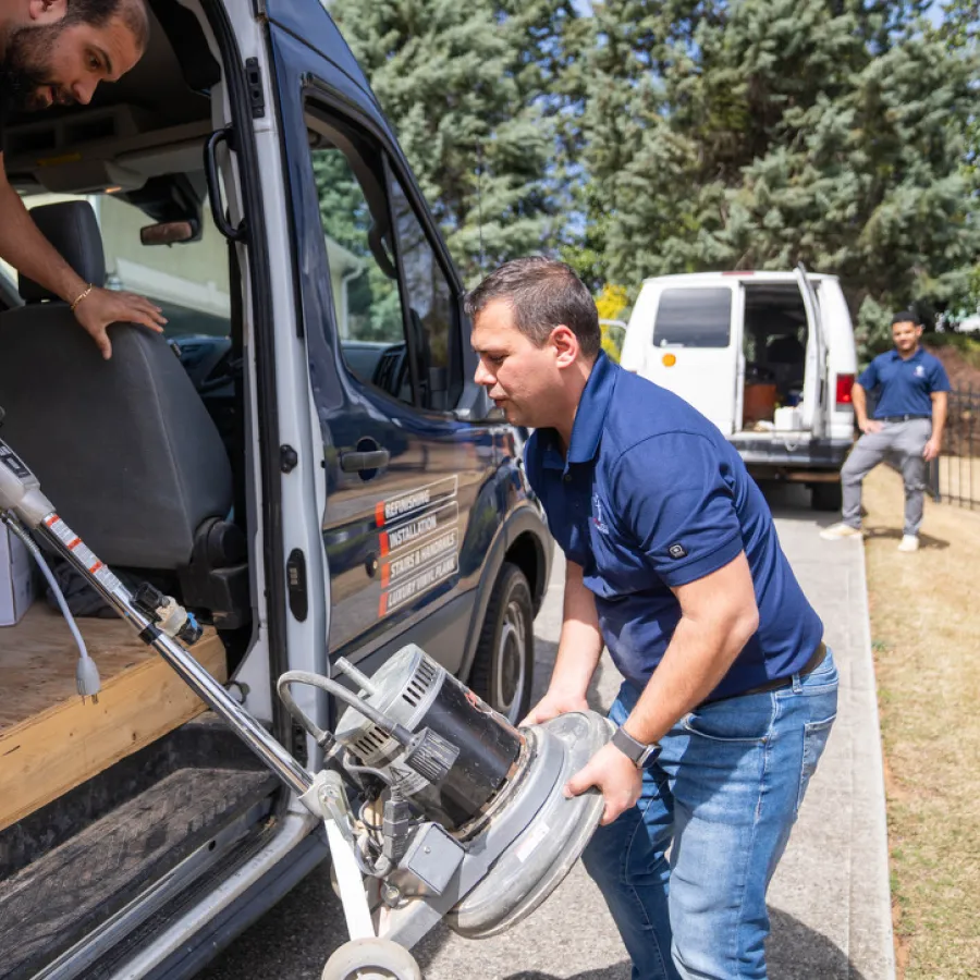 Two men loading or unloading a floor cleaning machine from a blue work van on a sunny day outdoors