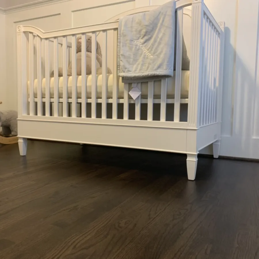 White baby crib with soft blanket and elephant plush toy on dark wood floor in cozy nursery room.