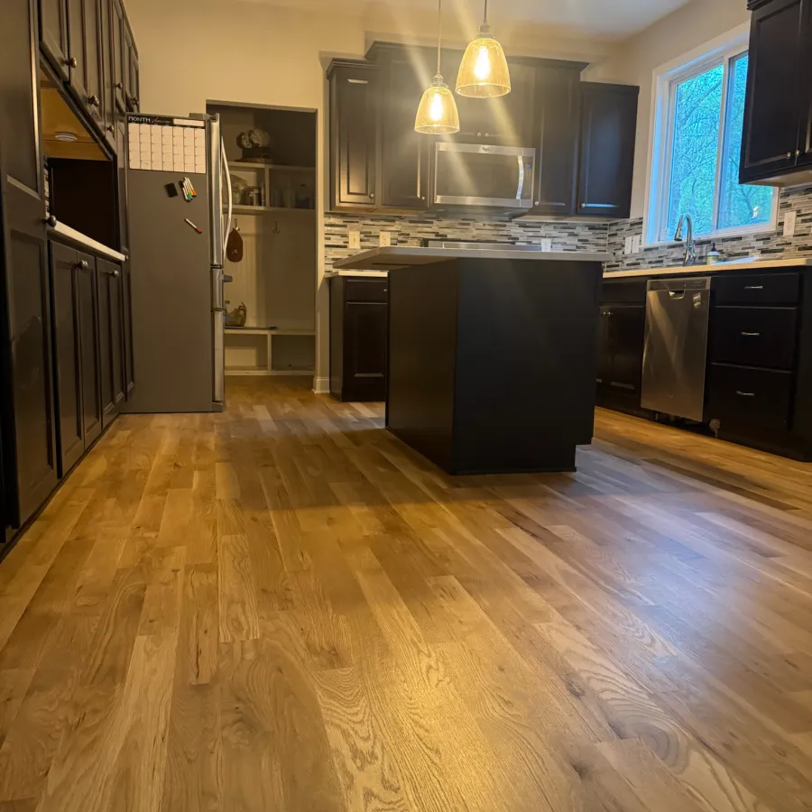 Modern kitchen with dark cabinets, wooden floor, central island, pendant lights, and stainless steel appliances.