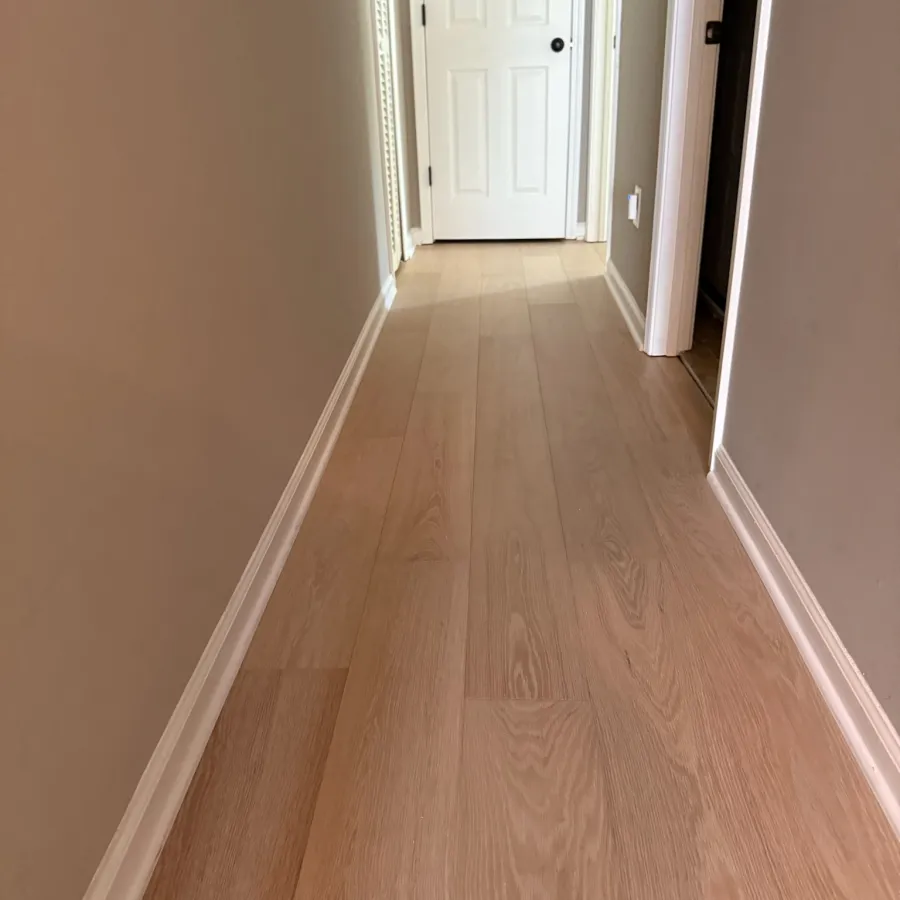 Narrow hallway with light wood flooring, beige walls, white baseboards, and a closed white door at the end.