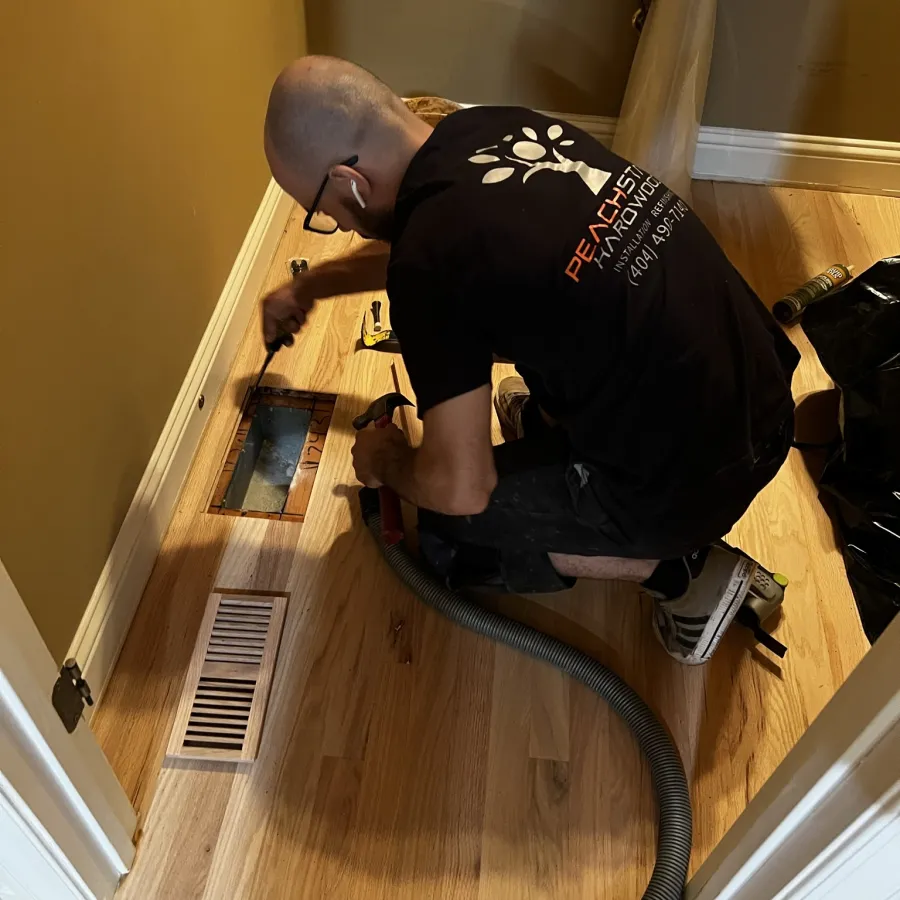 Worker installing hardwood flooring around vent in small bathroom with tools and vacuum hose nearby