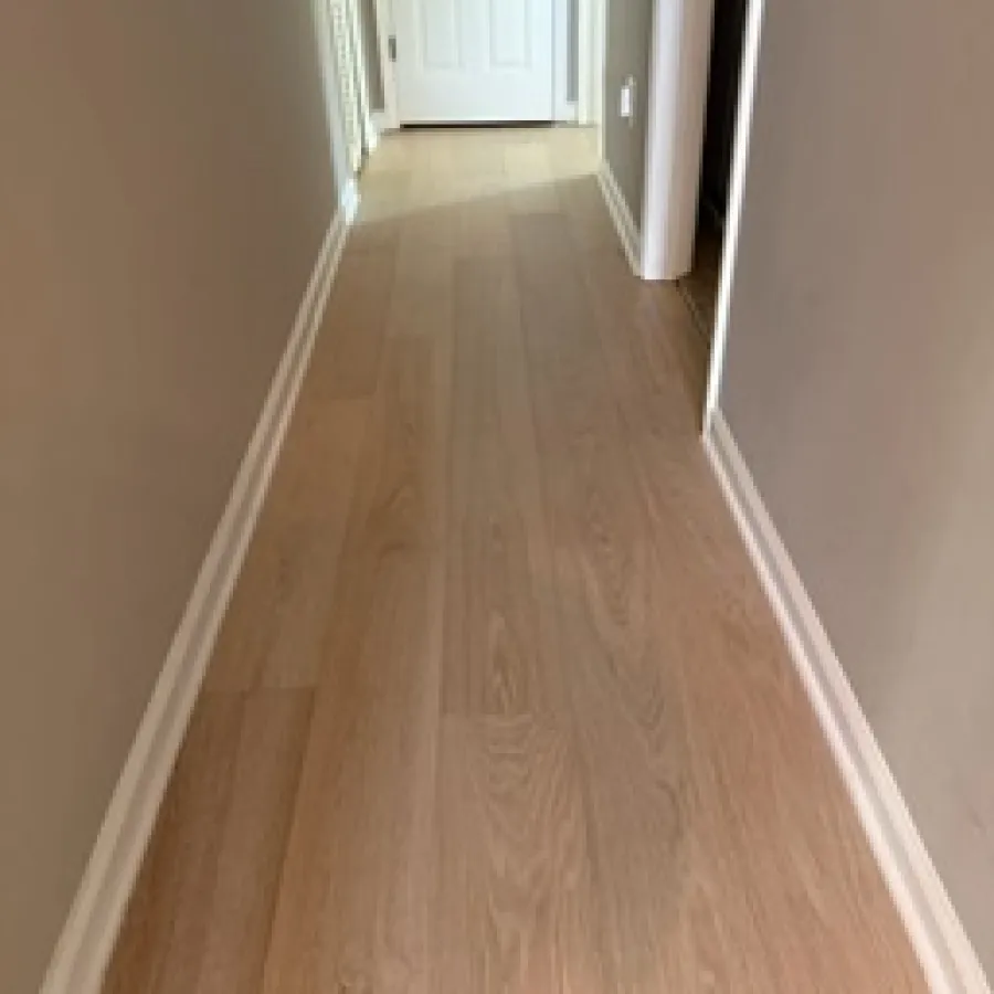 Narrow hallway with light wood flooring and beige walls leading to a white closed door at the end.