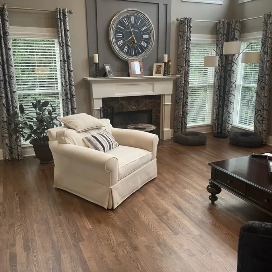 Cozy living room with beige armchair, large clock above fireplace, patterned curtains, and wooden floor.