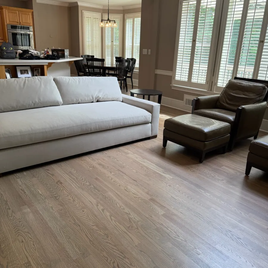 Bright living room with white sofa, brown leather armchair, wooden flooring, and large windows with shutters.