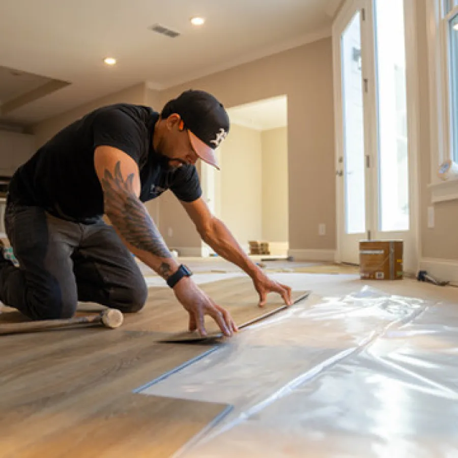 Man installing vinyl flooring in a room, measuring and laying out sheets carefully over wooden subfloor.