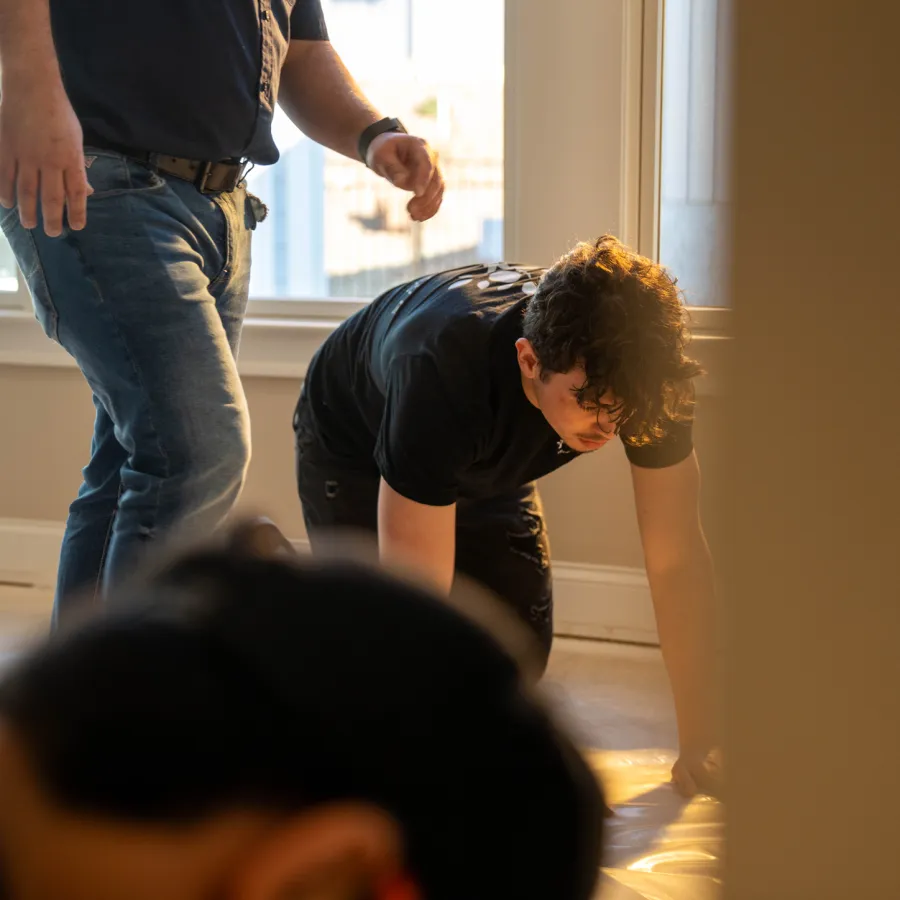 Two people working on the floor near a window with sunlight, one kneeling and the other standing.
