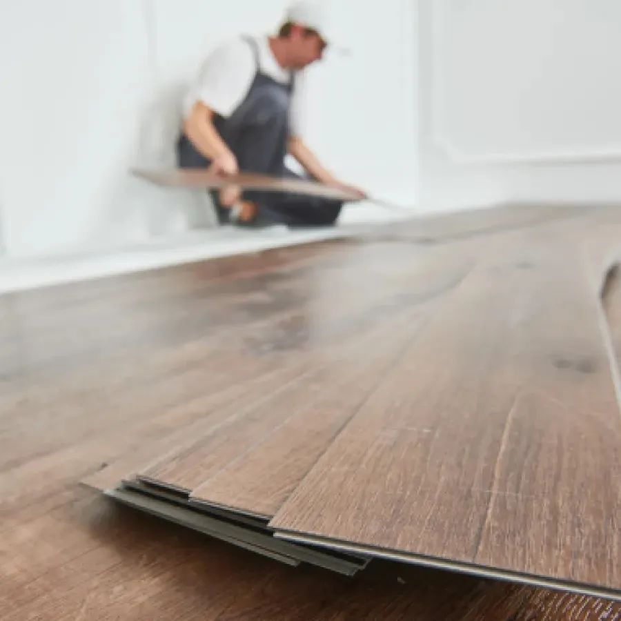Worker installing dark wood laminate flooring panels in a bright room with white walls.