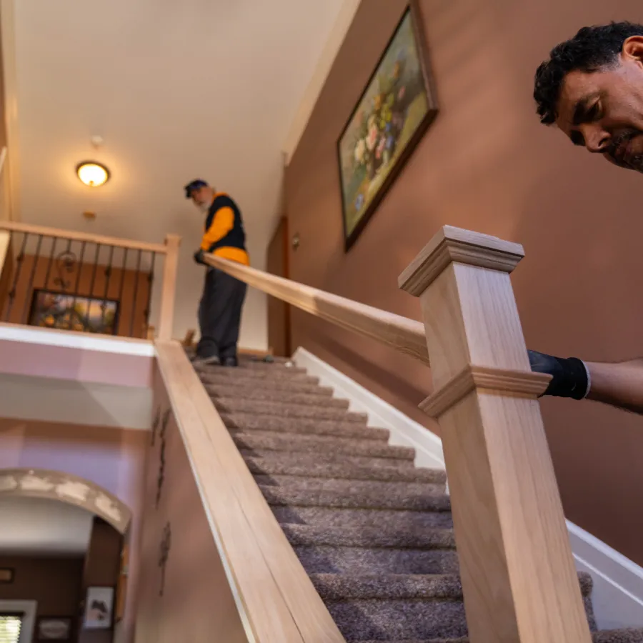 Two workers installing a wooden handrail on a carpeted staircase inside a home with brown walls and wall art.