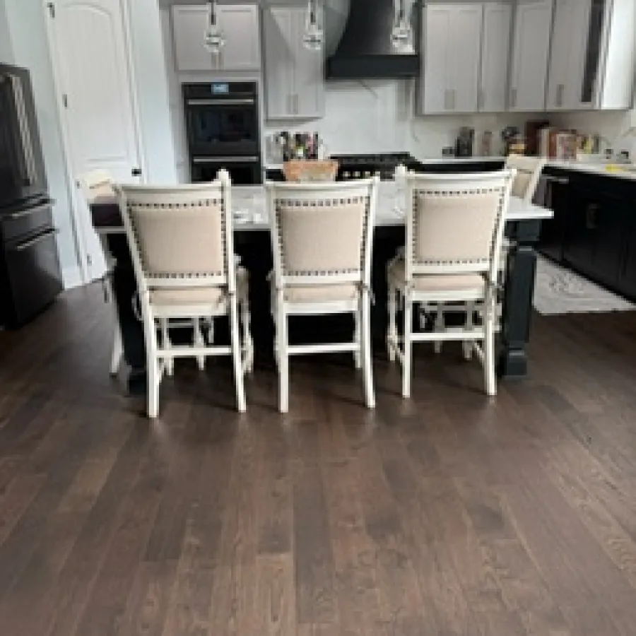 Modern kitchen with dark hardwood floors, white island with beige chairs, black appliances, and pendant lights.